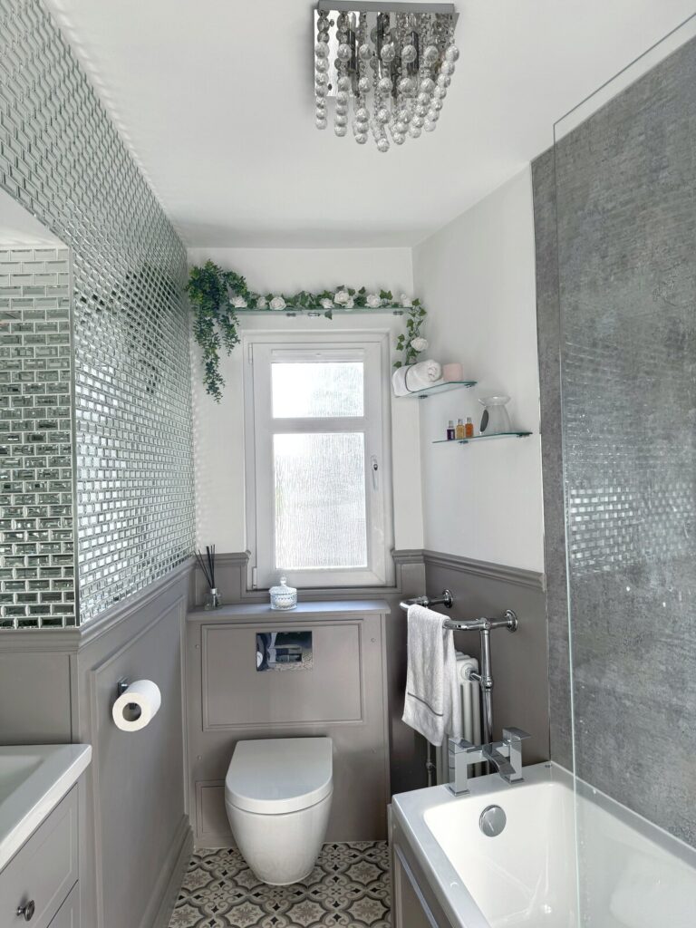 Stylish and elegant greige bathroom with textured wet wall, along with a full wall of mirrored tiles above the panelling. Modern vanity unit and patterned flooring and modern chandelier. Bathroom renovation after image.