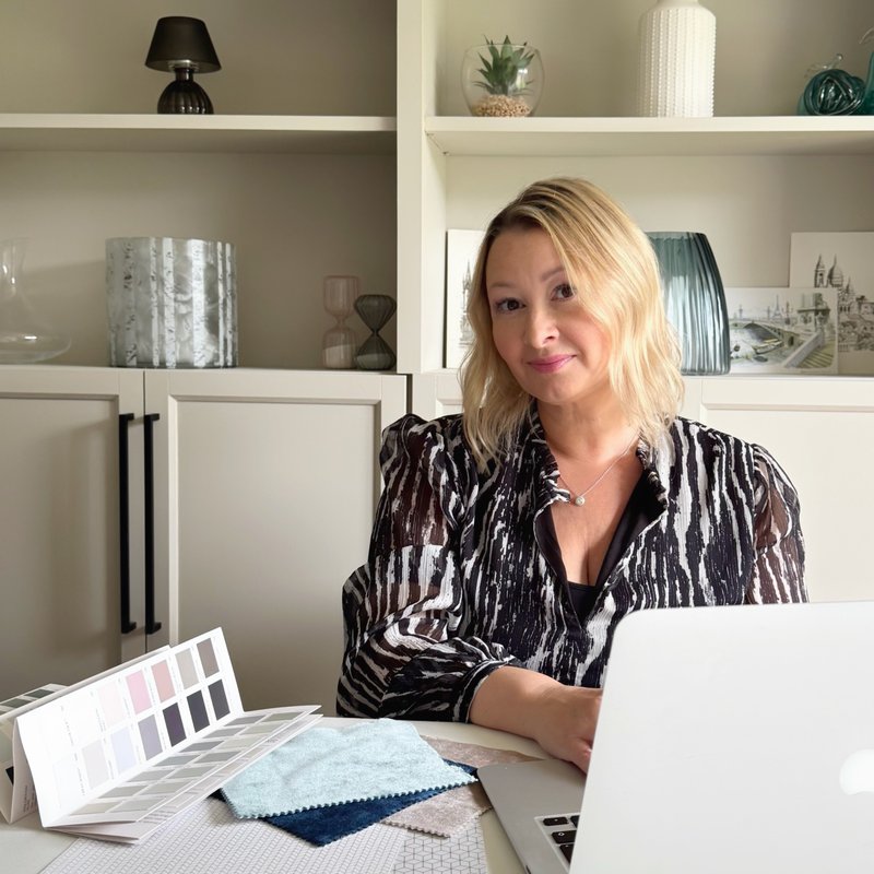 Image of Owner of Secret Home Designer home decor blog, sitting at work desk with fabric samples and laptop, with greige storage unit with home accessories in background.
