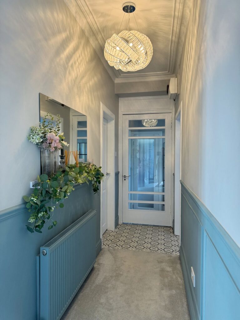 Modern hallway after renovation with a statement chandelier, panelled walls, and shelving with decorative accessories. Renovation after image.