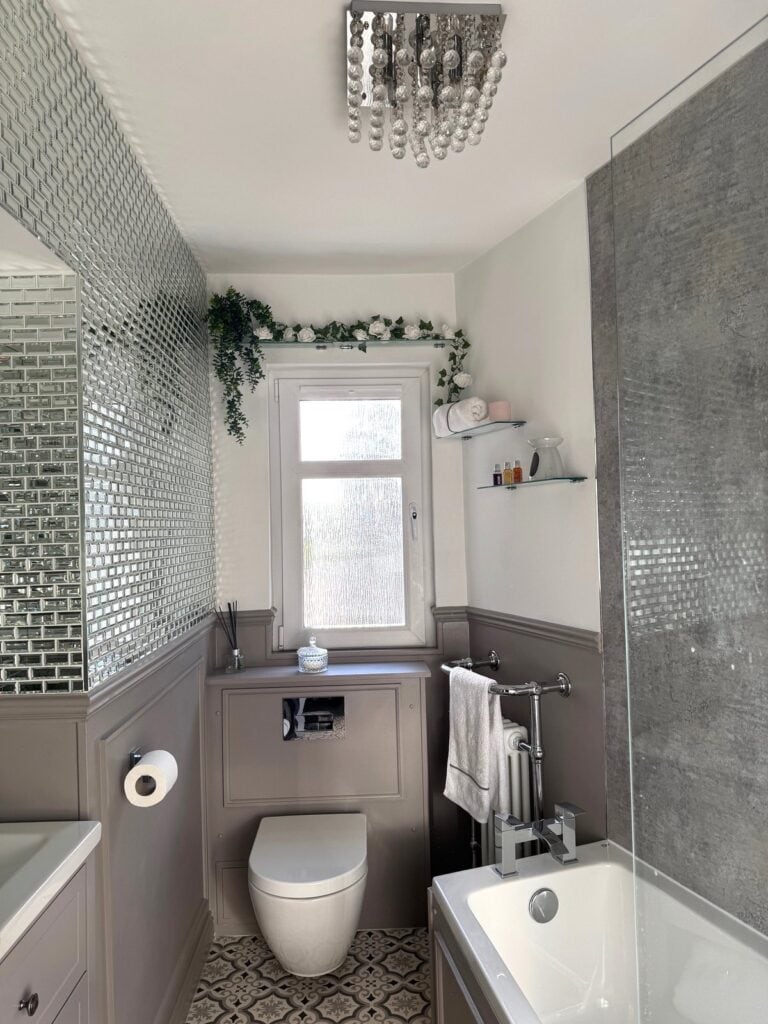 Stylish and elegant greige bathroom with textured wet wall, along with a full wall of mirrored tiles above the panelling. Modern vanity unit and patterned flooring and modern chandelier. Bathroom renovation after image.