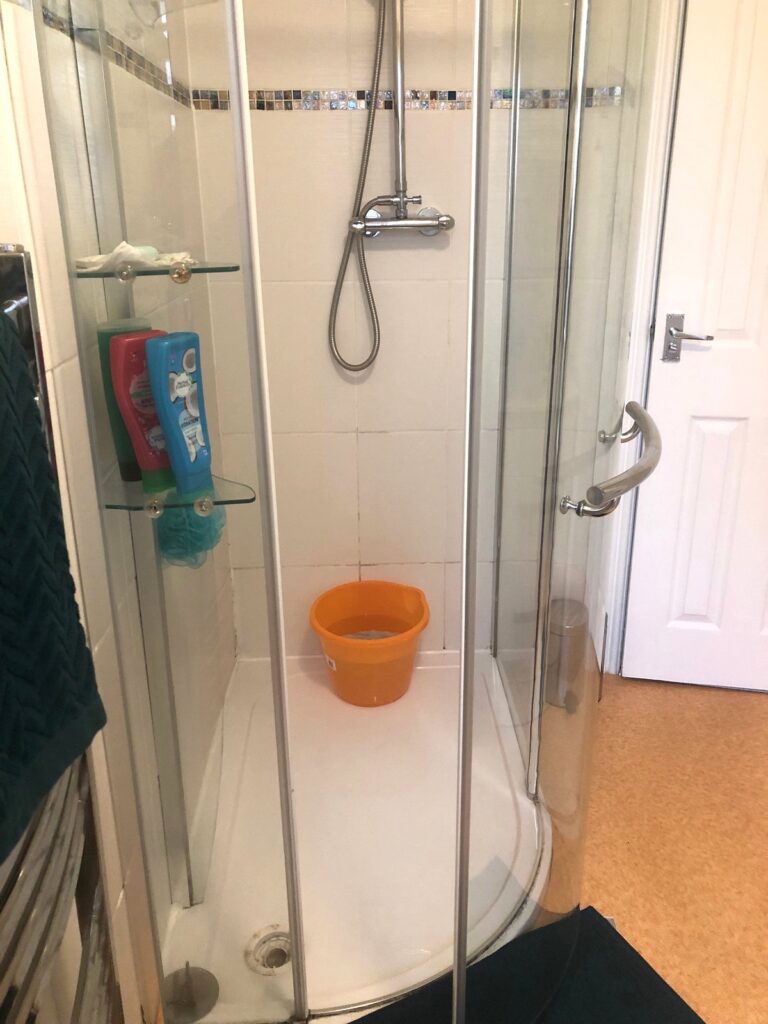 Photo of an outdated shower cubicle with glass doors and basic white tiles and dated orange vinyl. Before renovation image.