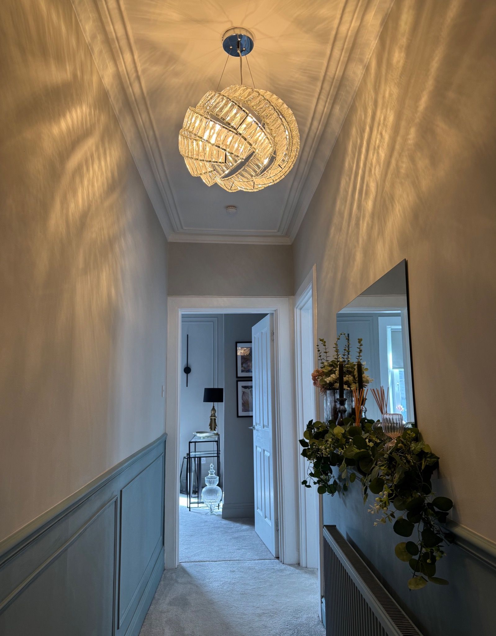 Modern hallway after renovation with a statement chandelier, panelled walls, and shelving with decorative accessories. Renovation after image.