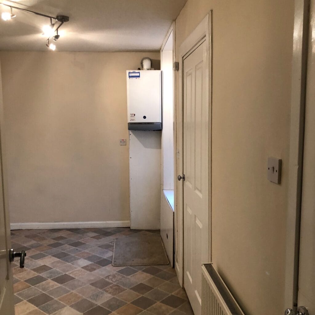 Outdated kitchen corner before renovation with beige walls, vinyl flooring, and exposed boiler. Before renovation image.
