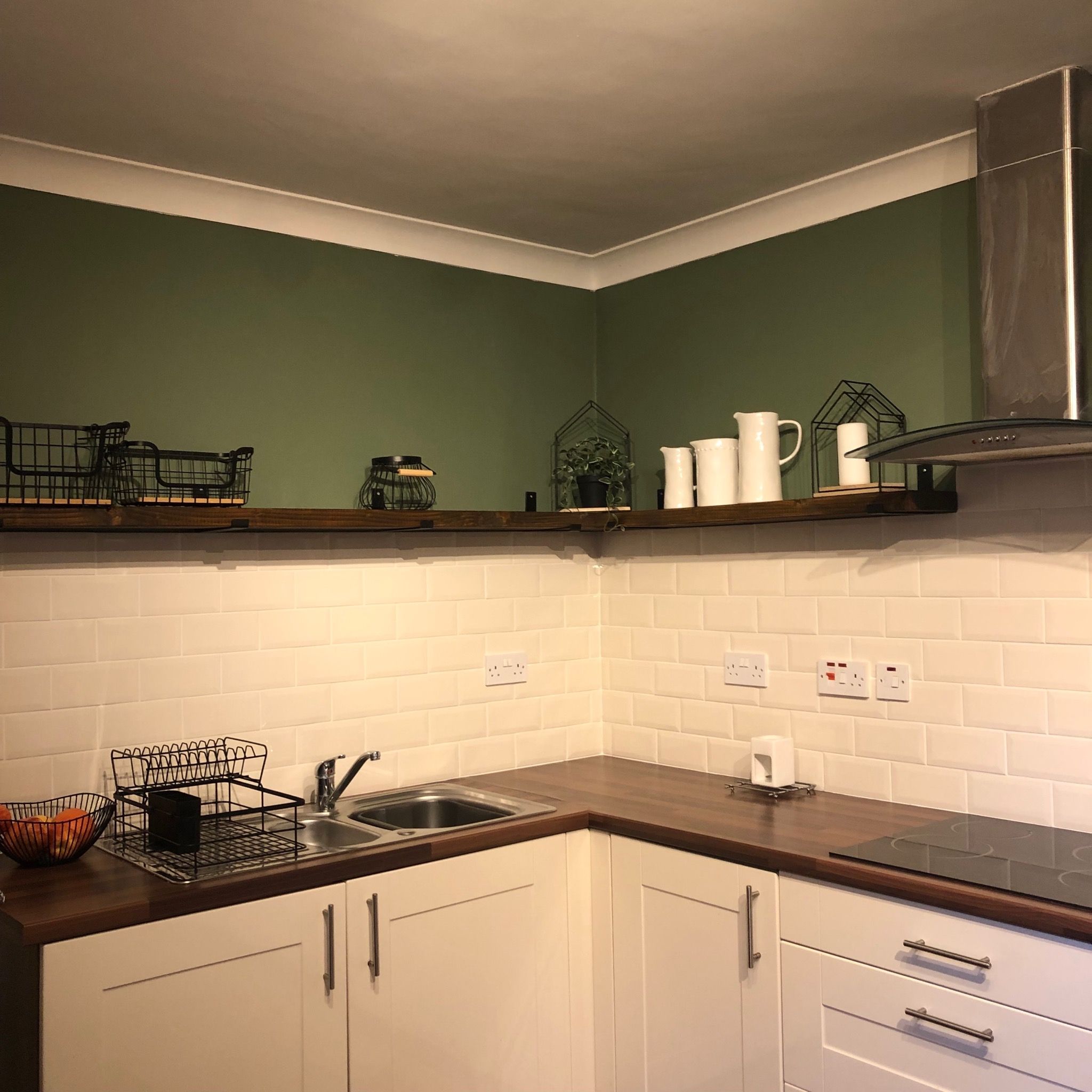 Modernized kitchen with dark green painted walls, open shelving, cream subway tile backsplash, open shelving and wood-effect countertop. After renovation image.