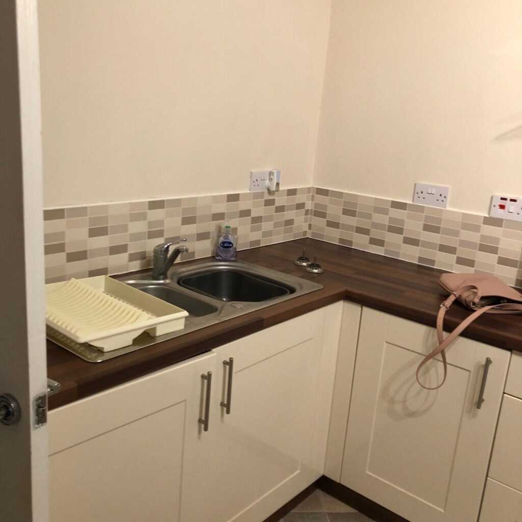Traditional kitchen area before renovation with beige mosaic tile backsplash and double stainless steel sink. Before renovation image.