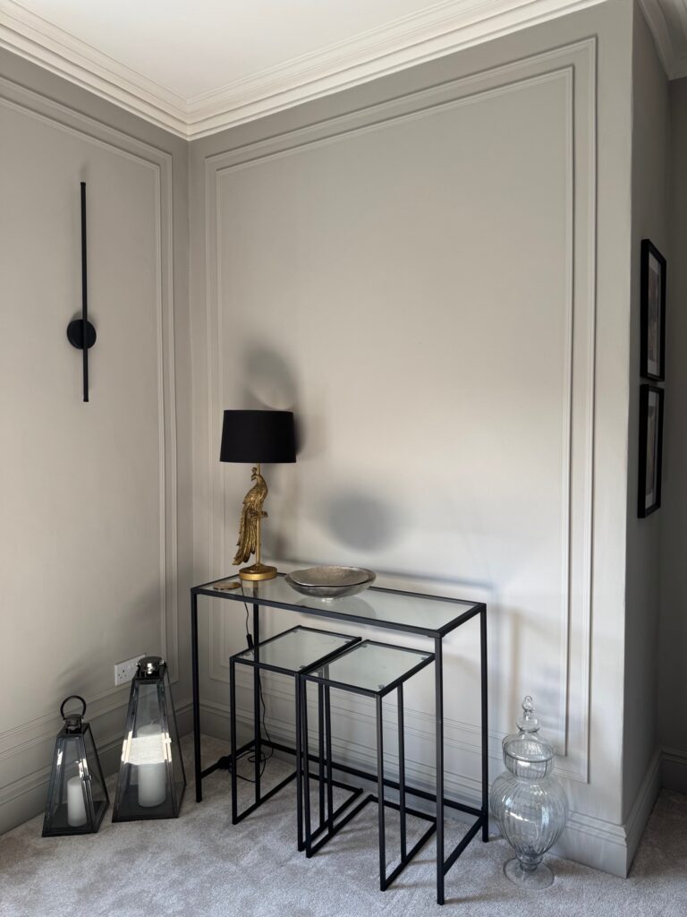 Contemporary corner of a styled living room with glass console tables, gold lamp, black details and greige wall panelling. After renovation image.