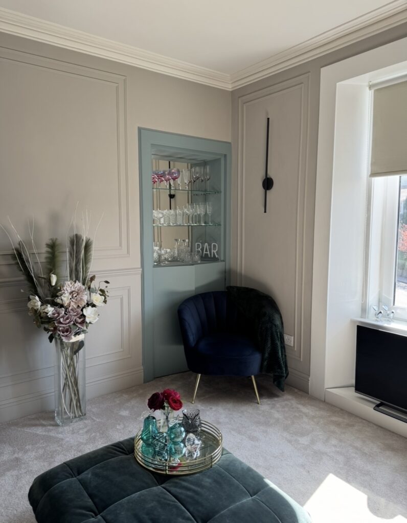 Stylish, modern living room with greige wall panelling, display bar area, and navy velvet armchair beside a large window. After renovation image.