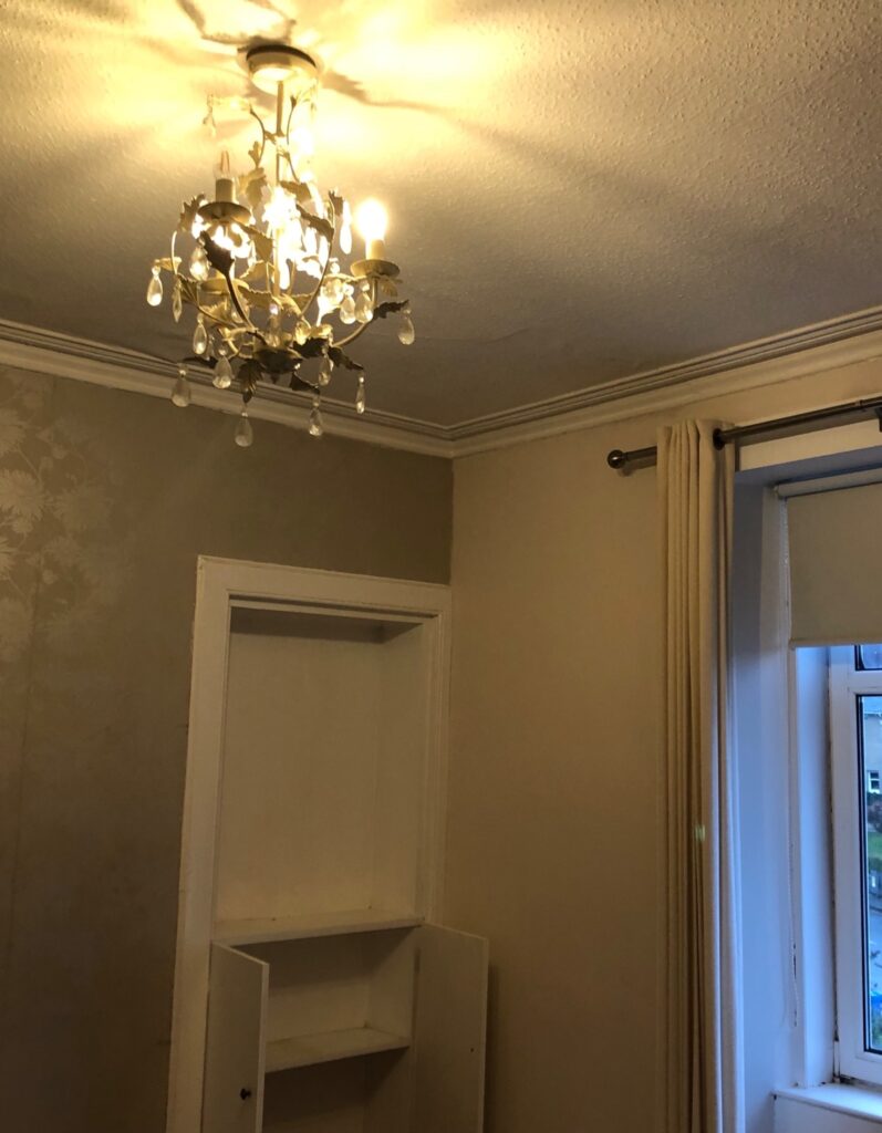 Before image of living room corner with old chandelier, old fitted shelves, and window area. Before renovation image.