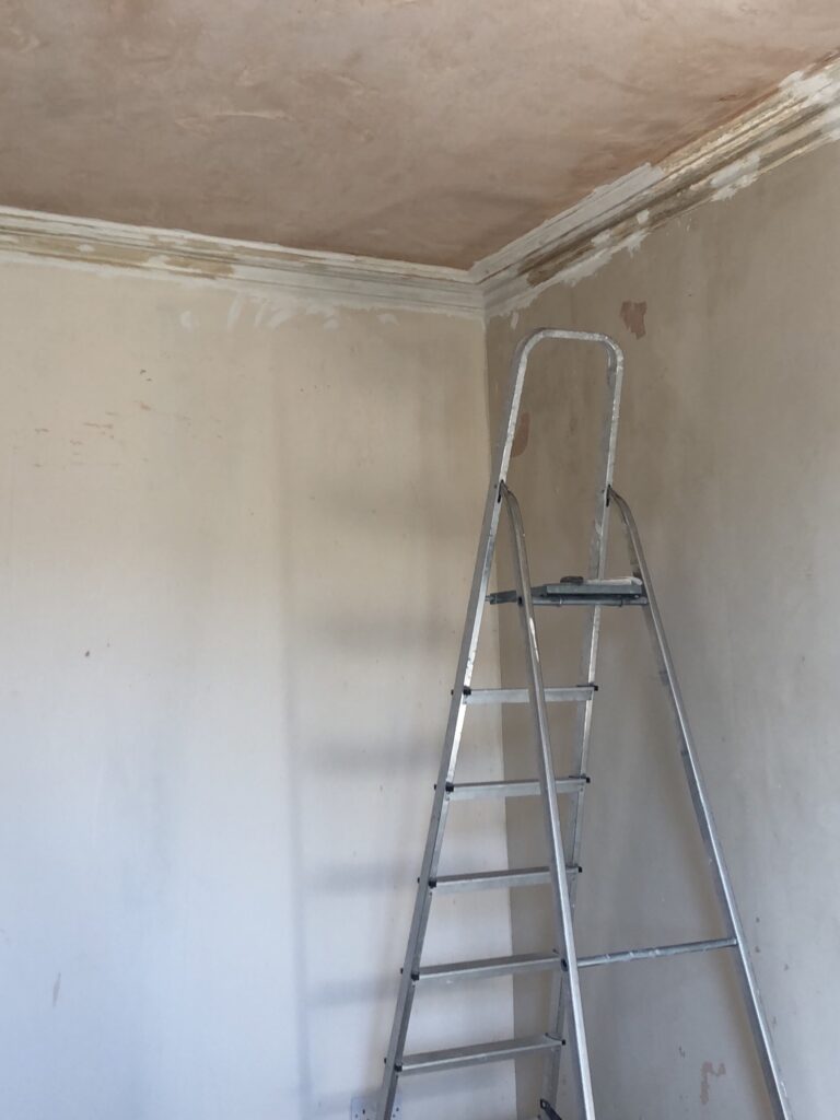 Corner of a living room. Coving is being repaired, mid-renovation with bare plaster ceiling, ladder, and patchy walls. Living room before renovation image.