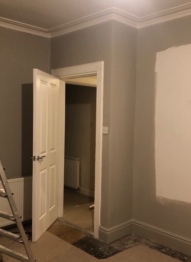 Partially painted living room wall mid-renovation with greige paint and exposed flooring.