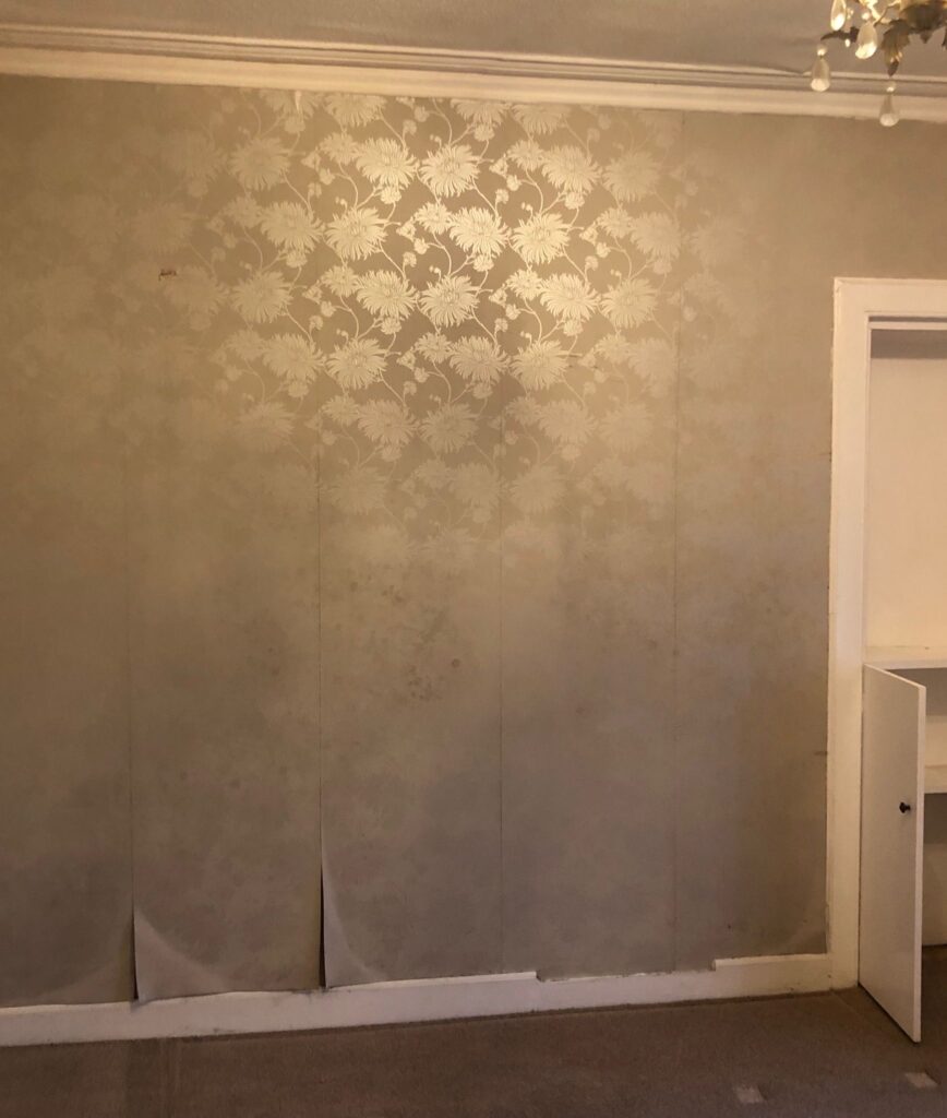 Living room wall before renovation with outdated wallpaper peeling at the bottom and faded floral pattern.