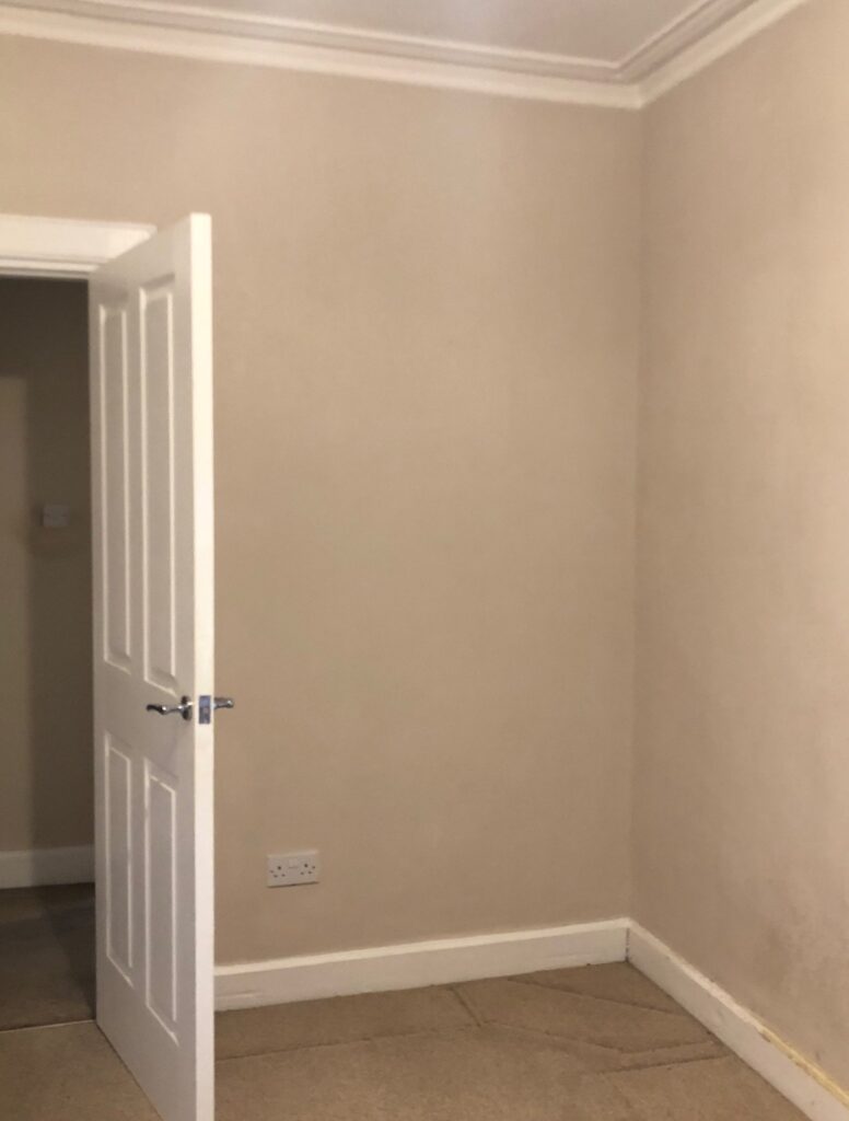 Empty corner of a spare room before renovation, showing beige walls and carpet.